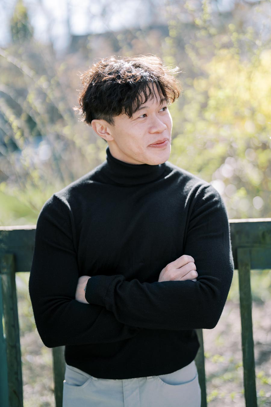 A person stands outdoors with arms crossed, wearing a black turtleneck sweater, and looking to the side.
