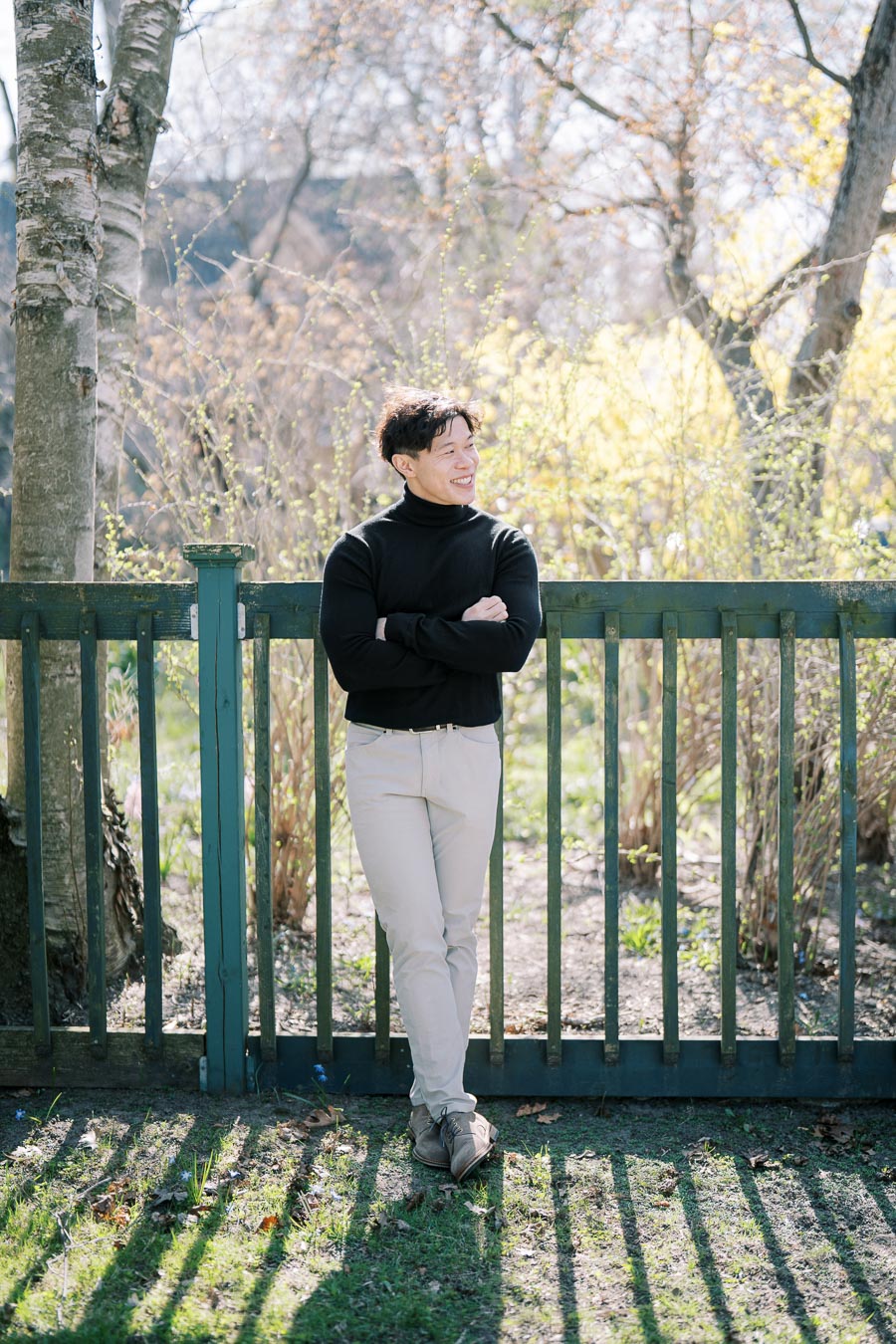 A person stands casually against a wooden fence, enjoying a sunny day in a park-like setting.