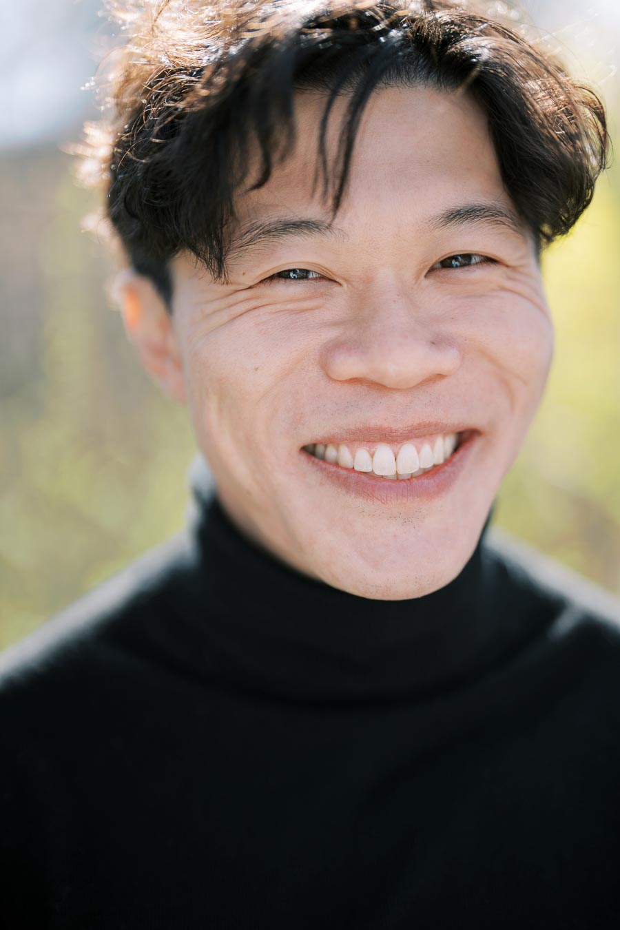 A person smiling warmly in an outdoor setting, wearing a black turtleneck.