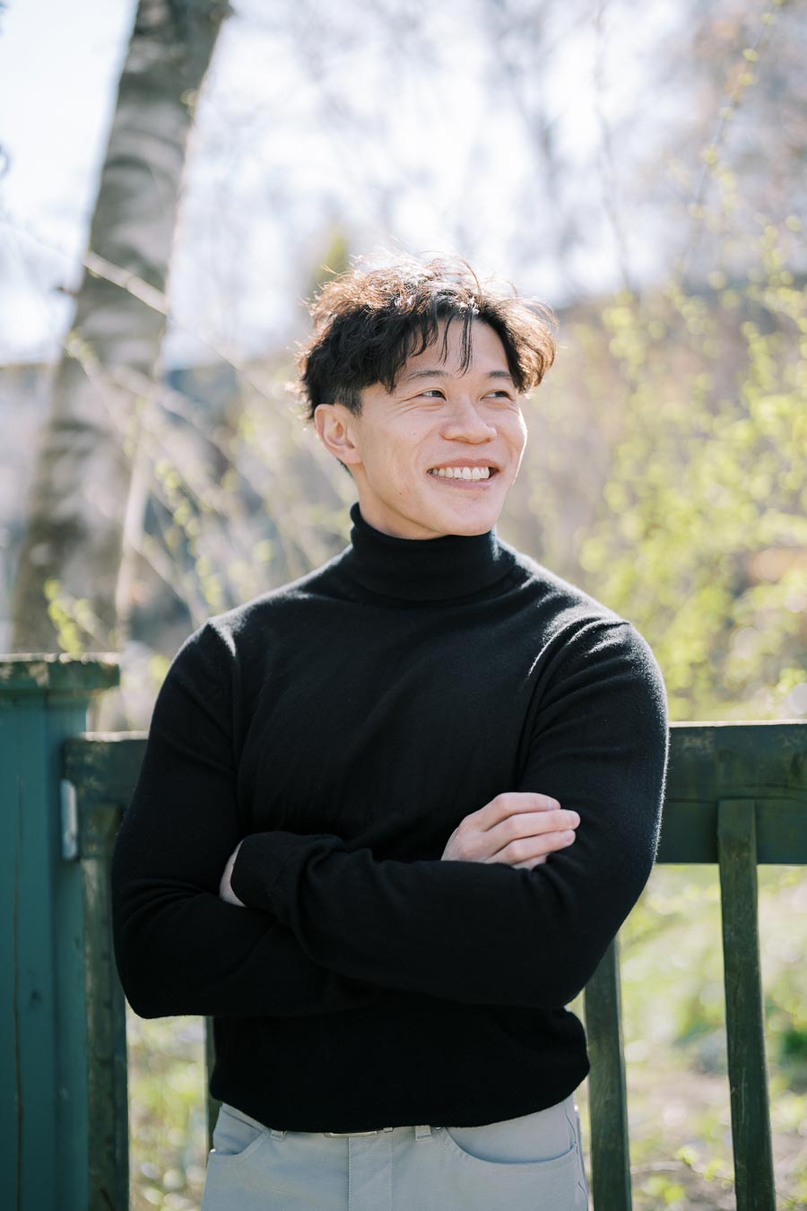 A person in a black turtleneck smiles while standing outdoors, surrounded by trees and greenery.
