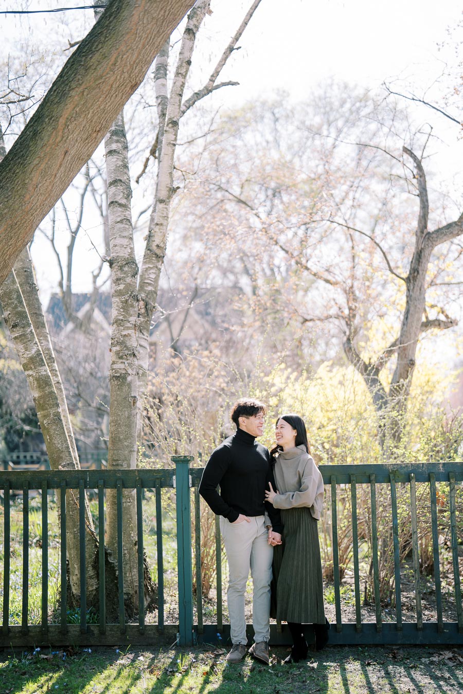 A couple enjoys a sunny day in a park, standing by a wooden fence surrounded by trees.