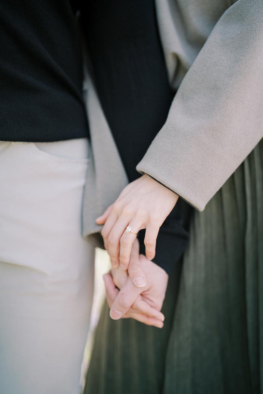 A close-up image of a couple holding hands, highlighting a woman's engagement ring. The couple is dressed in neutral tones, with the woman's hand gently resting on the man's.