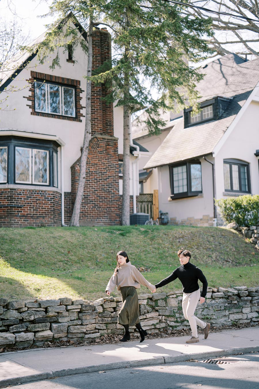A couple enjoys a sunny day, walking hand in hand past a charming brick and stucco house.