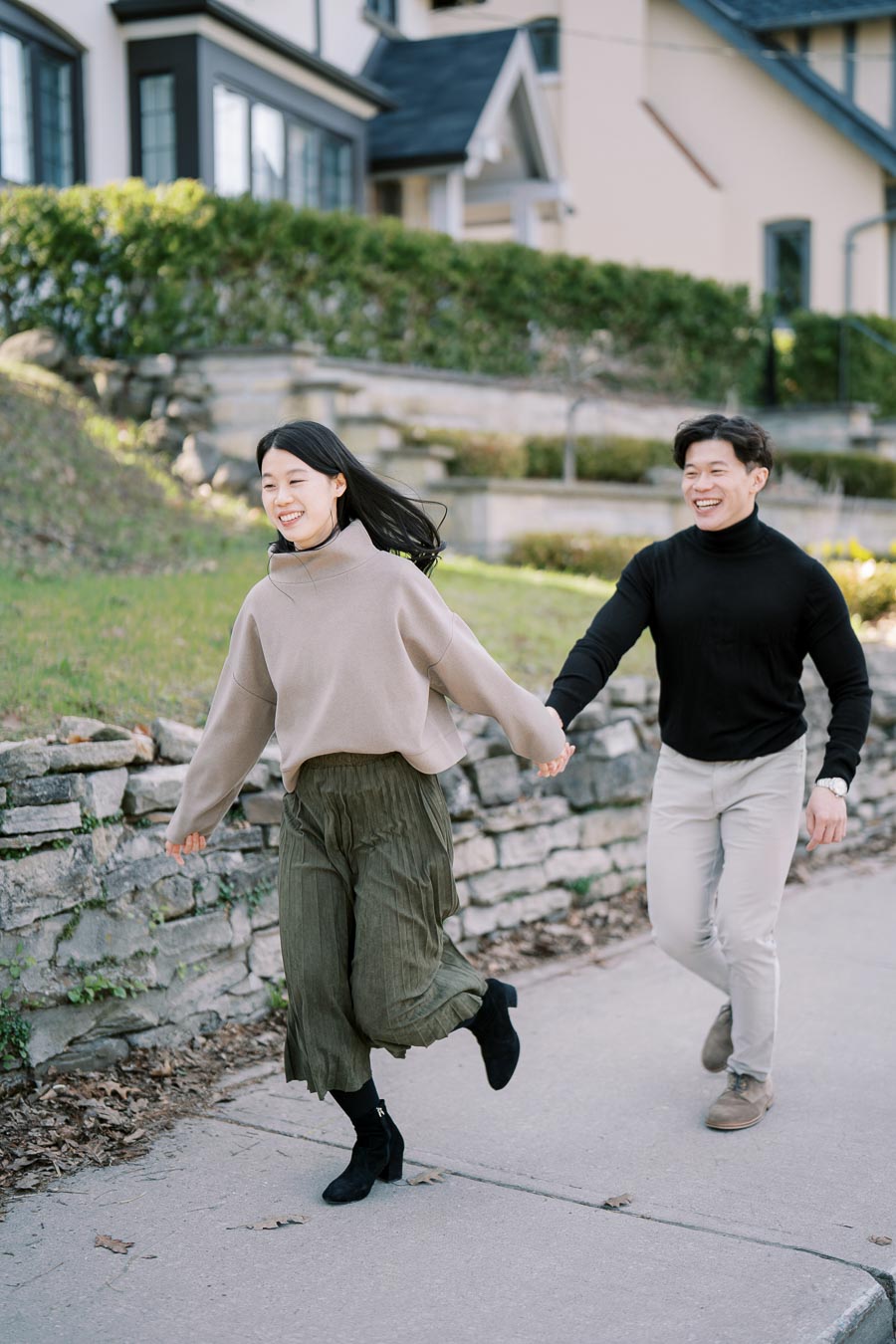 A couple joyfully runs hand in hand along a sidewalk in a residential area, surrounded by stone walls and greenery.