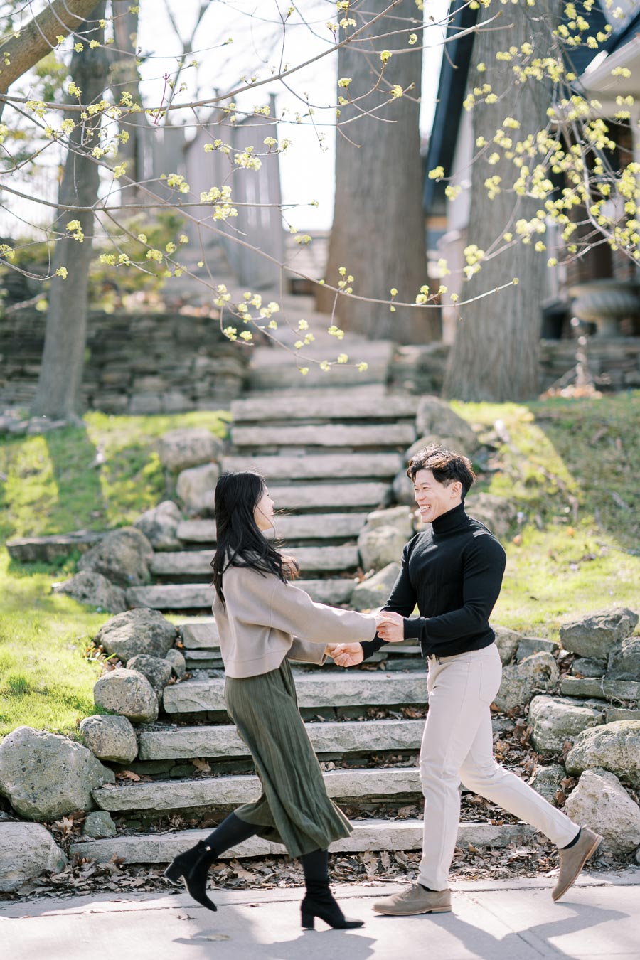 A couple joyfully holds hands in a park setting, surrounded by stone steps and budding trees, capturing a moment of happiness and connection.