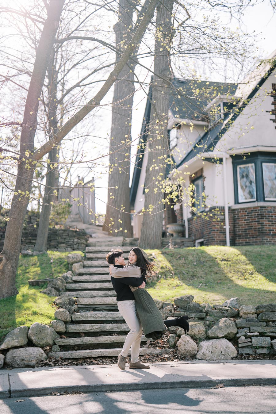 A joyful couple embraces in front of a charming house with stone steps and budding trees, capturing a moment of happiness and springtime warmth.