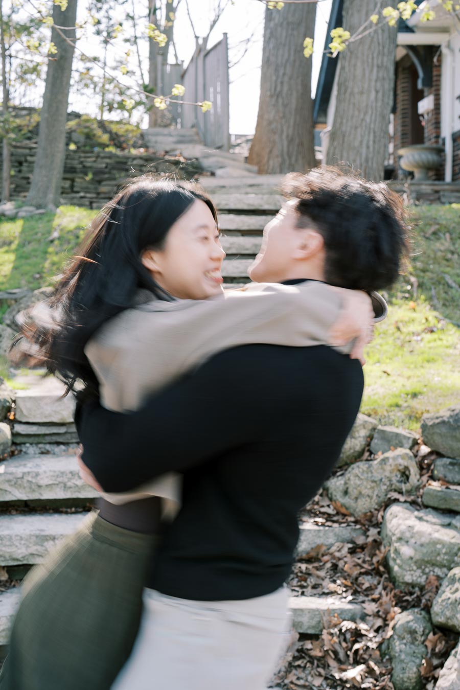 A joyful couple embraces and spins on a stone pathway in a wooded area, capturing a moment of happiness and motion.