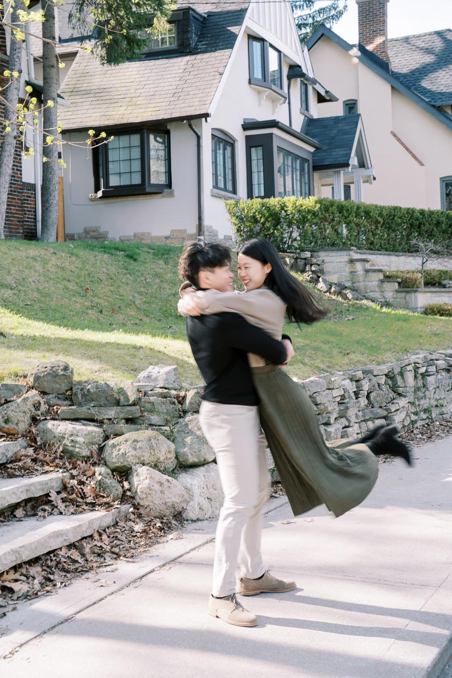 A joyful couple embraces and spins on a sunny day in front of charming houses.