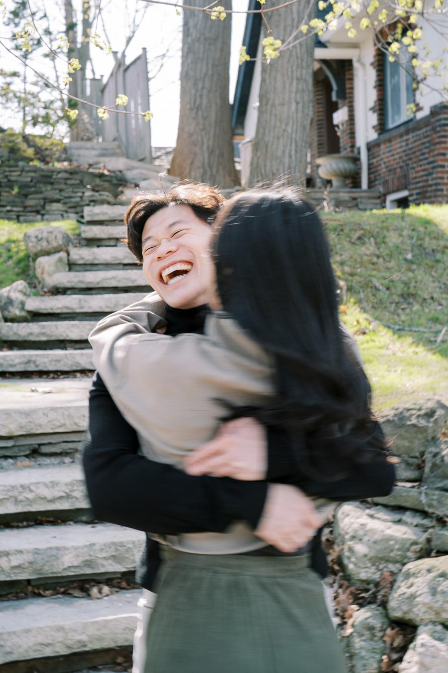 A couple shares a joyful embrace outdoors near stone steps and a house, surrounded by trees and greenery.