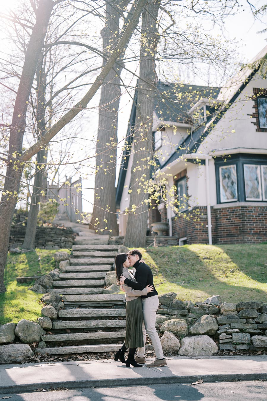 A couple shares a romantic kiss in front of a charming house with stone steps and budding trees, capturing a serene and intimate moment.