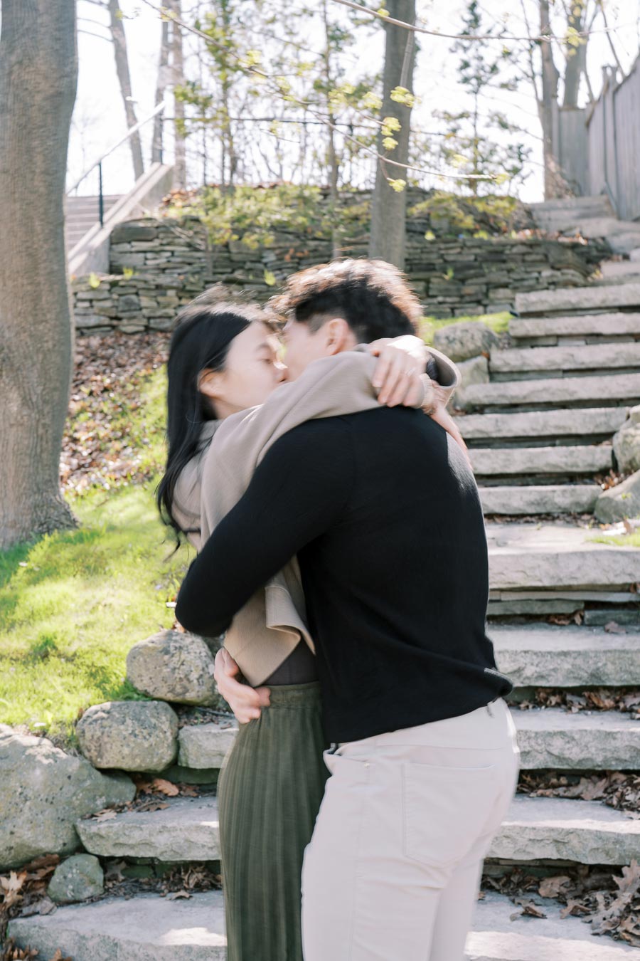 A couple shares a romantic moment, embracing and kissing near stone steps in a park setting.