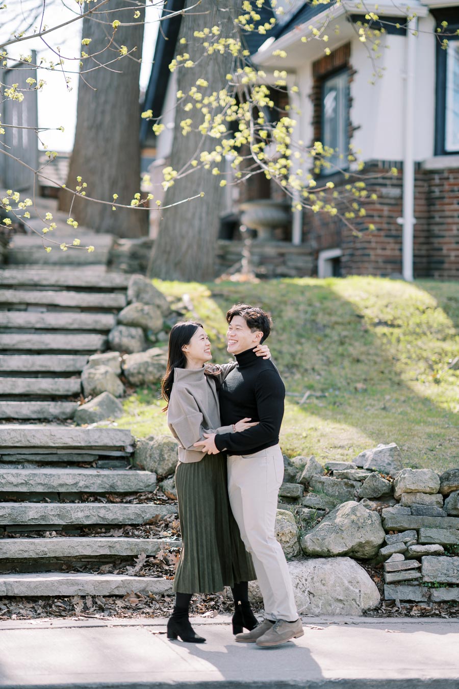 A couple shares a joyful moment in a garden setting, surrounded by stone steps and a charming brick house.