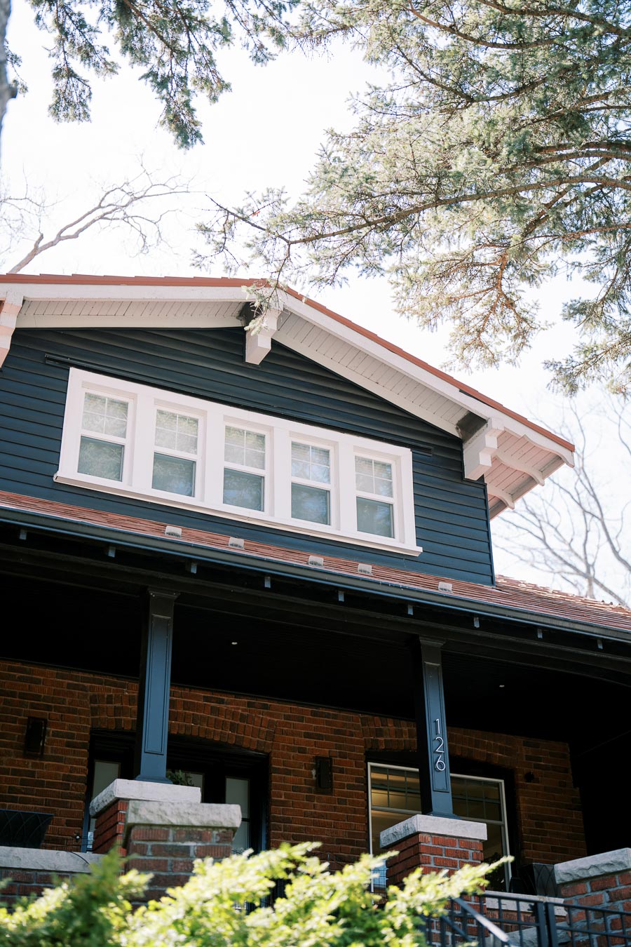 A charming two-story house with dark blue siding and white trim, featuring a brick lower level and a welcoming porch.
