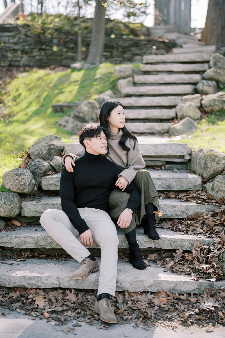 A couple enjoys a peaceful moment on stone steps in a scenic park setting, surrounded by greenery and fallen leaves.