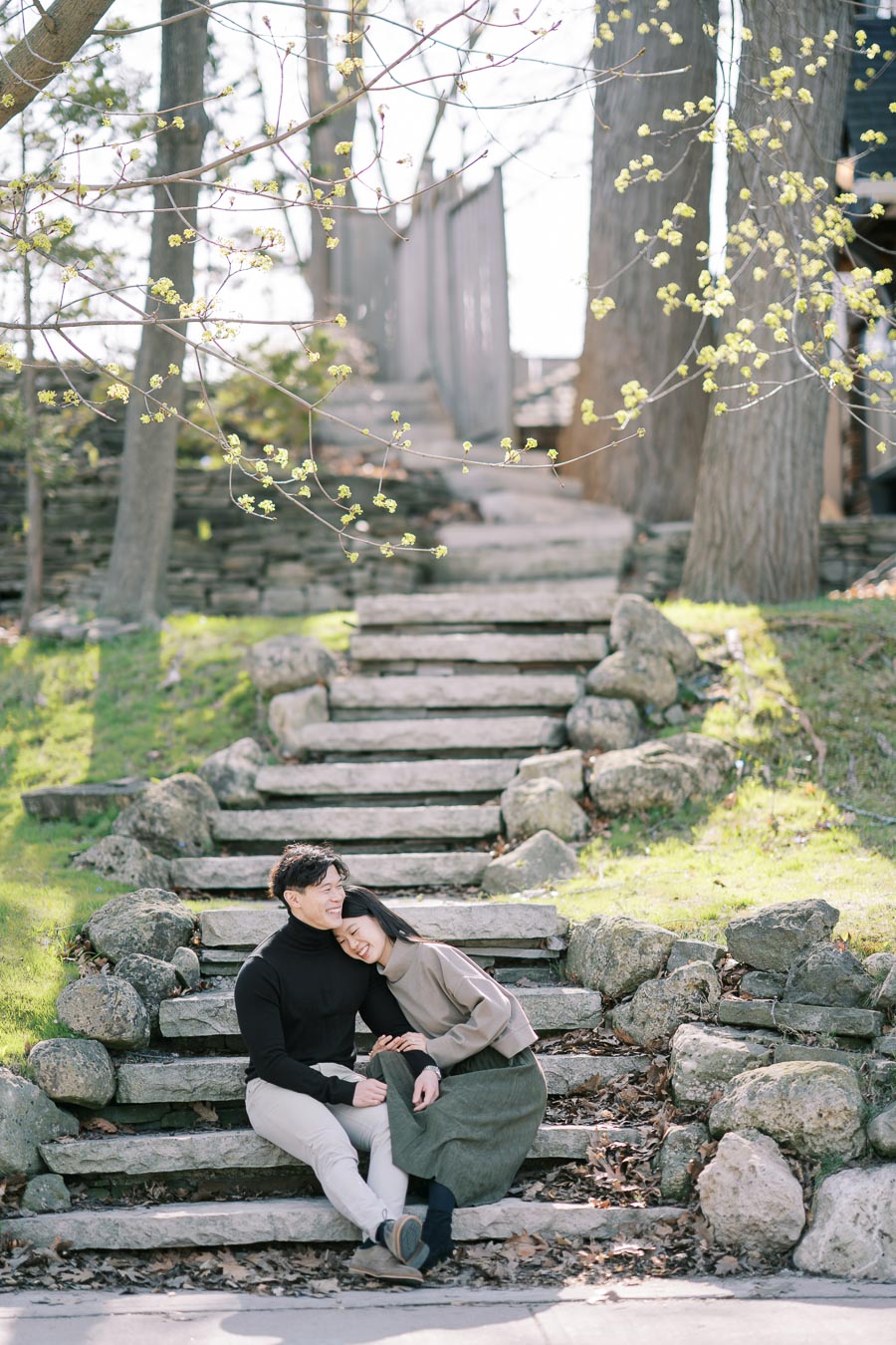 A couple enjoys a peaceful moment on stone steps amidst a serene park setting, with budding trees signaling the arrival of spring.