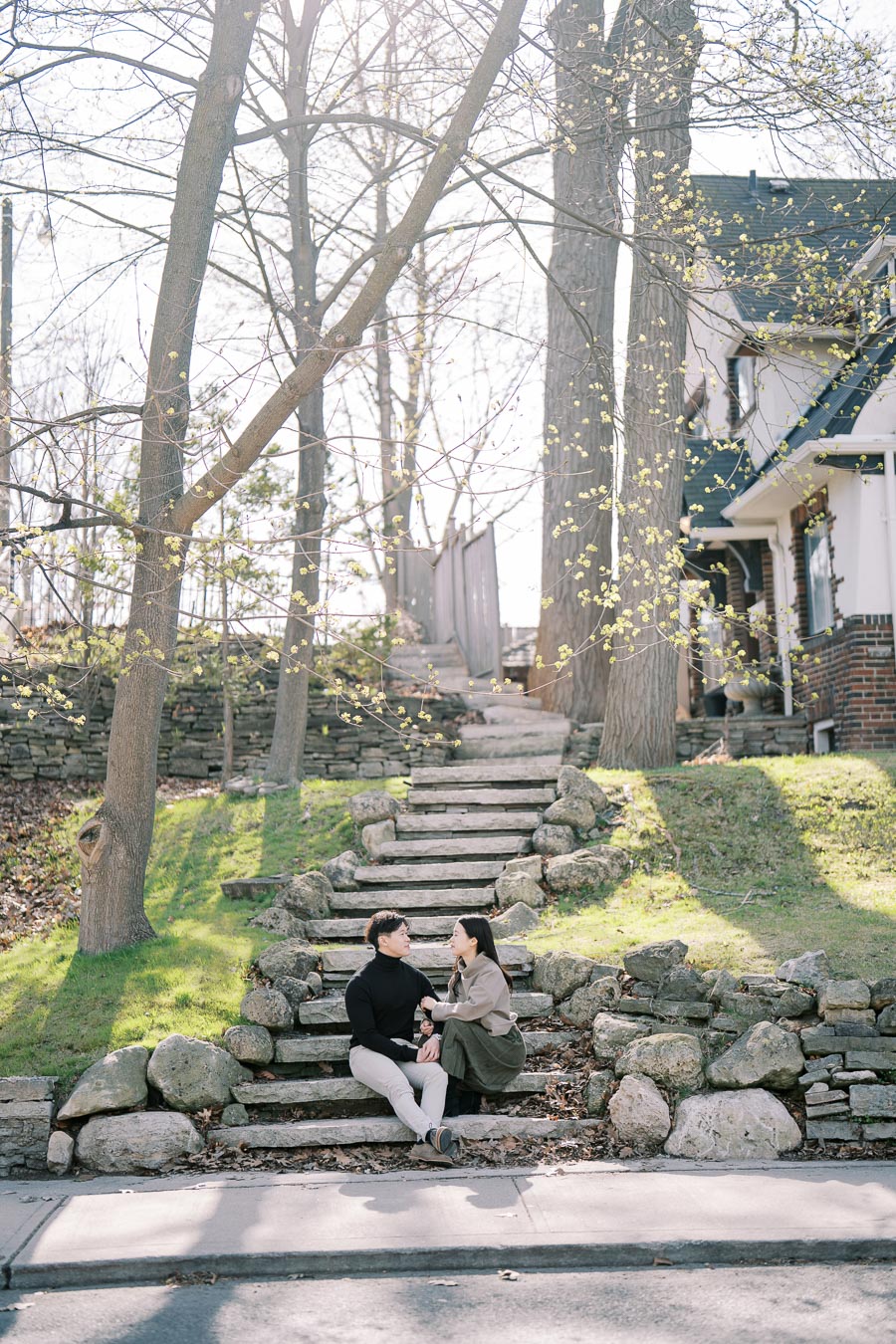 A couple sits on stone steps in a serene, tree-lined setting with a house in the background, enjoying a peaceful moment together.