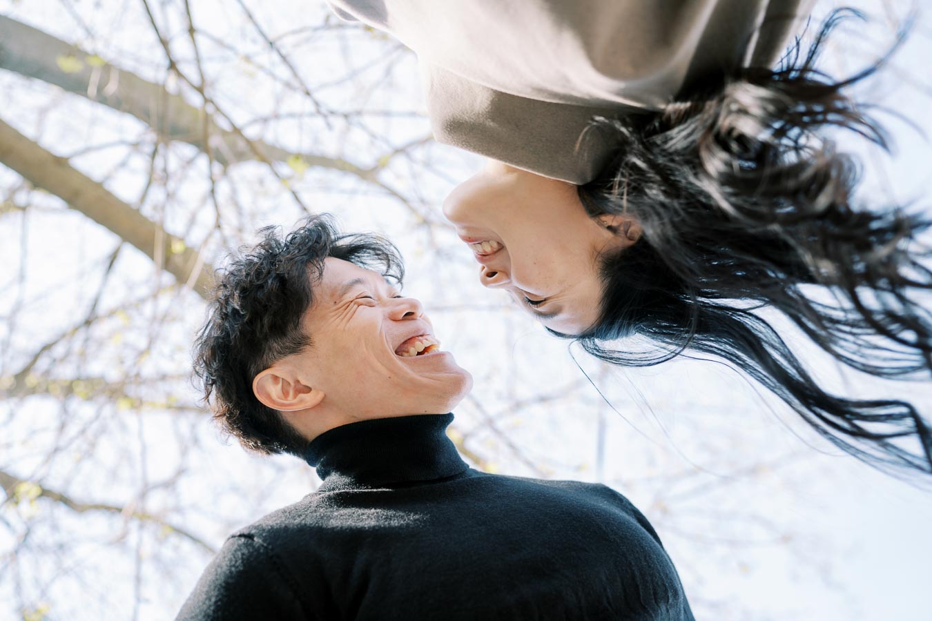 A couple shares a joyful moment, laughing together under a tree on a sunny day.