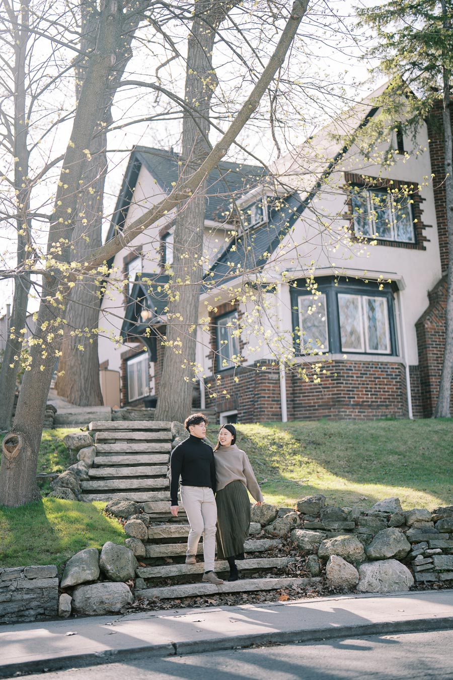 A couple enjoys a walk down stone steps in front of a charming Tudor-style house, surrounded by budding trees and springtime greenery.