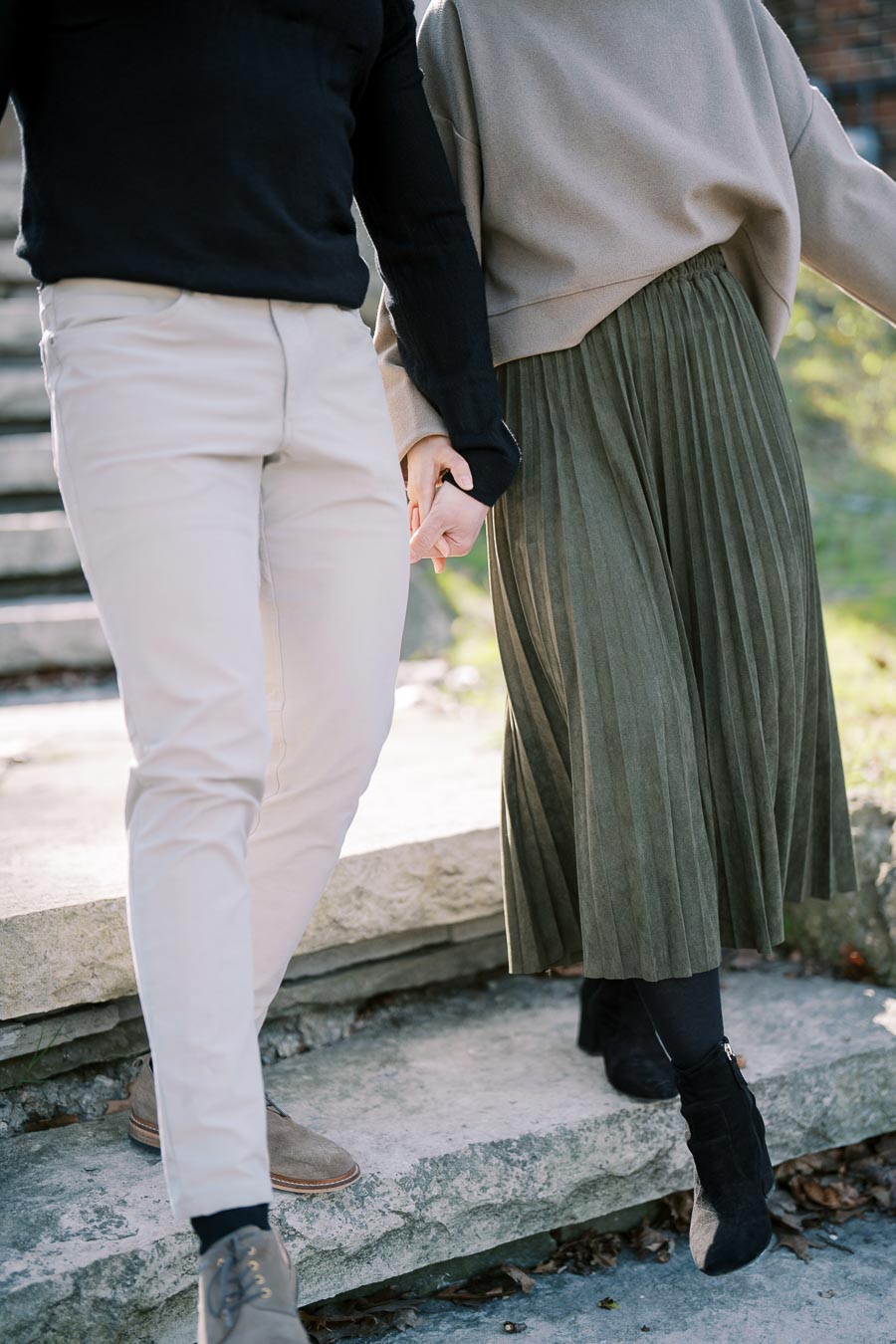 A couple holding hands while walking up stone steps, dressed in casual attire. The man is wearing beige pants and a black sweater, while the woman is in a pleated green skirt and beige sweater.