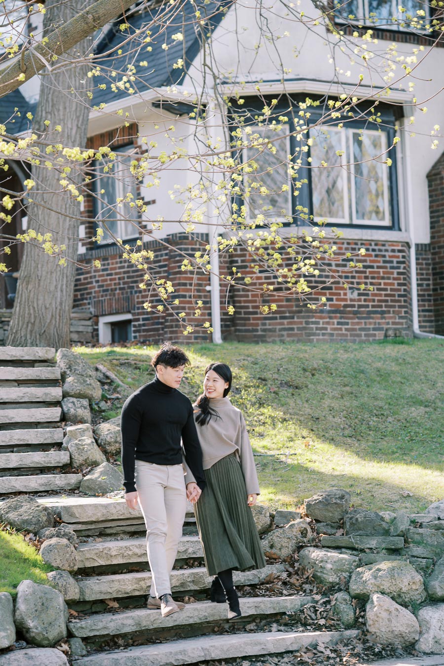 A couple enjoys a walk down stone steps in a garden, surrounded by budding trees and a charming brick house in the background.