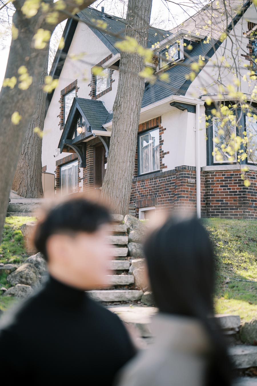 A Tudor-style house with a brick and stucco exterior is partially obscured by two blurred figures in the foreground, surrounded by trees and greenery.
