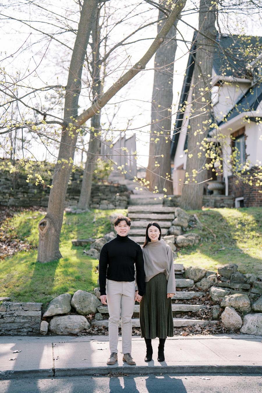 A couple stands on a sidewalk in front of a stone staircase leading to a house, surrounded by budding trees.