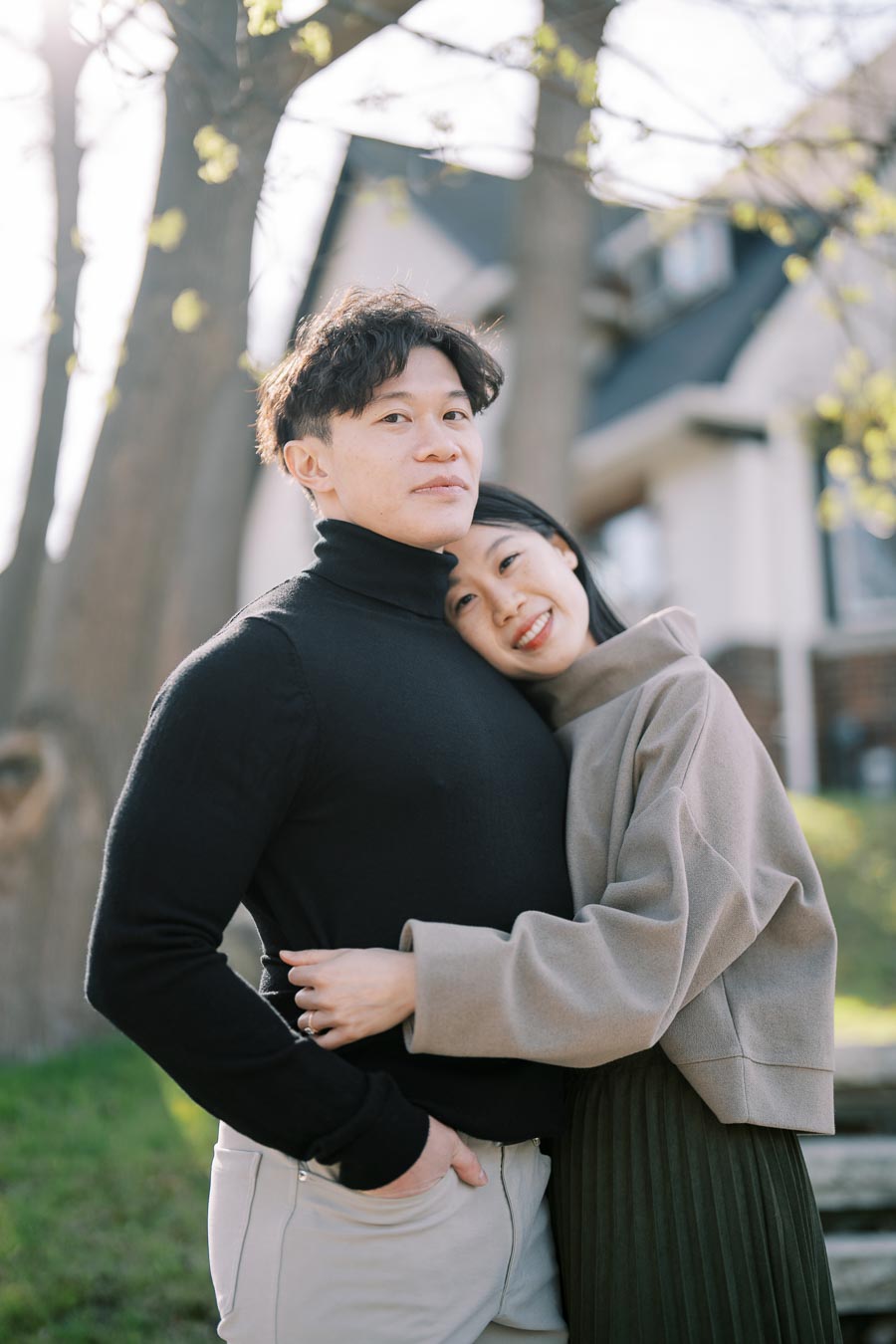 A couple embraces outdoors, with trees and a house in the background, capturing a moment of affection and warmth.