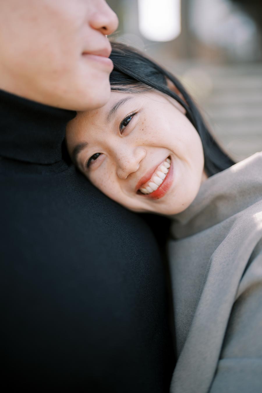 A woman smiles warmly as she rests her head on a companion's shoulder, capturing a moment of affection and happiness.
