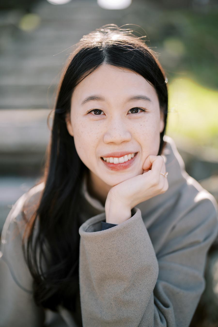 A woman smiling warmly in an outdoor setting, wearing a gray coat.
