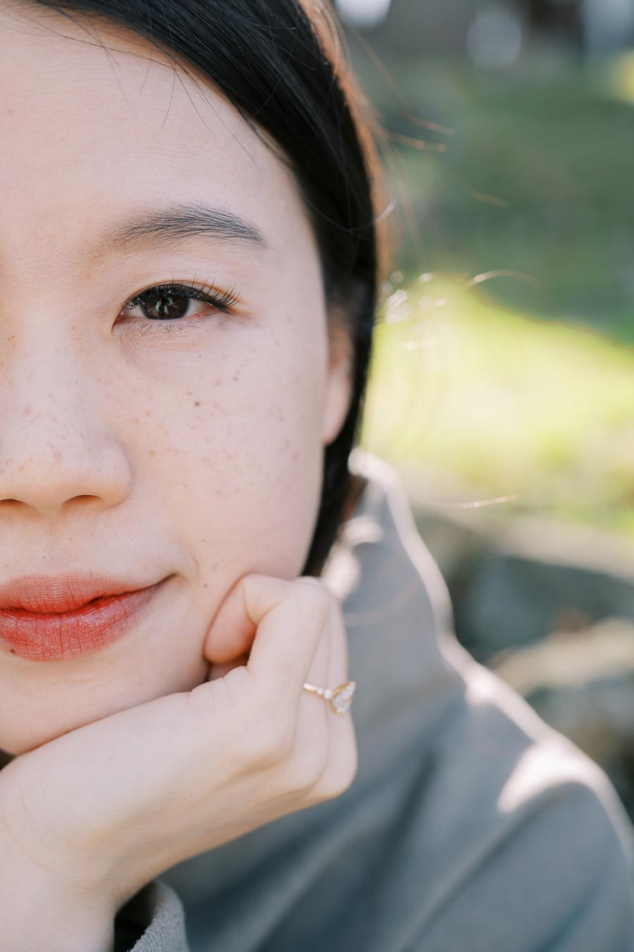 A close-up portrait of a woman with freckles, resting her chin on her hand. She is wearing a ring and appears to be outdoors, with a blurred green background.