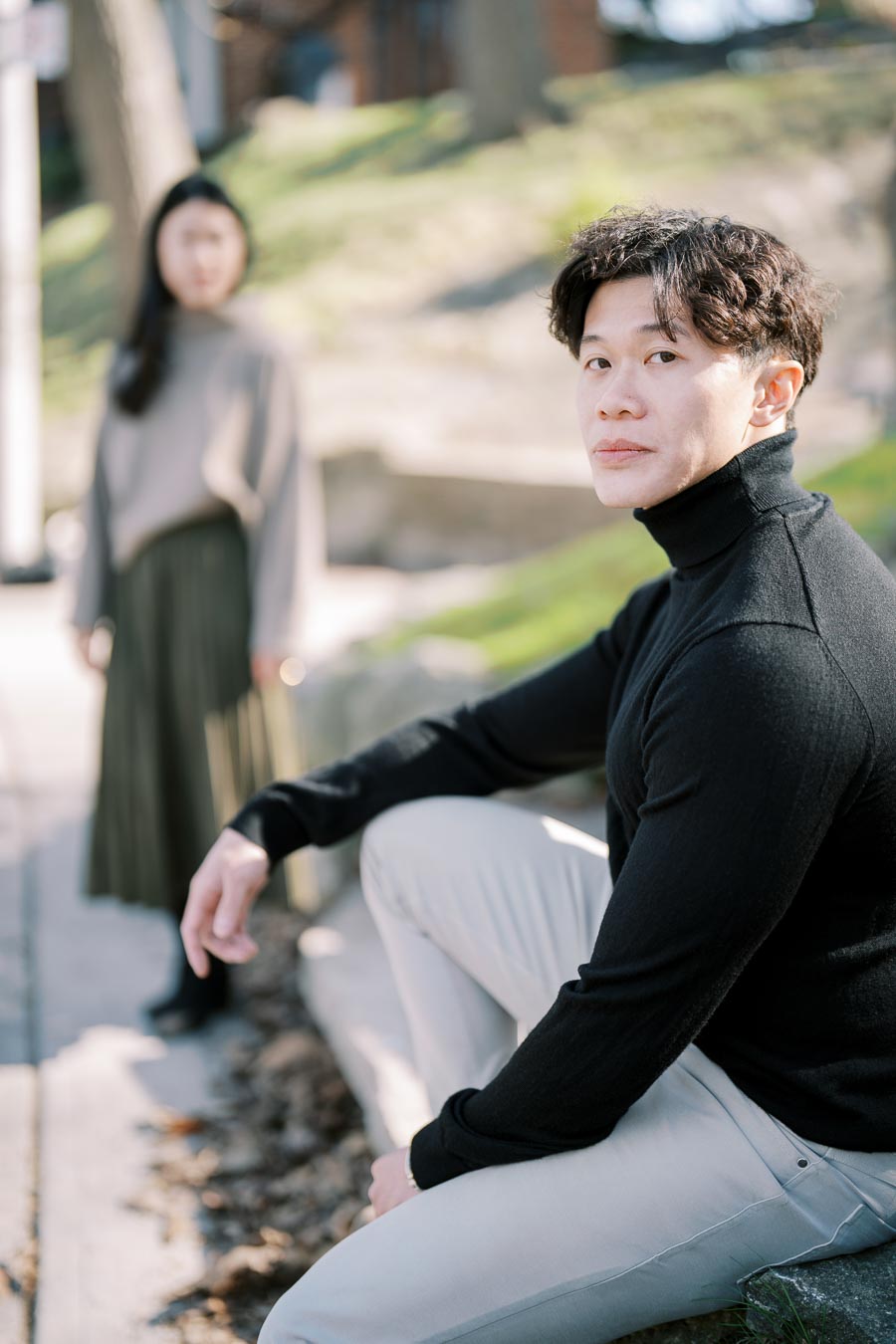 A man sits outdoors in a black turtleneck, while a woman stands in the background, slightly out of focus.
