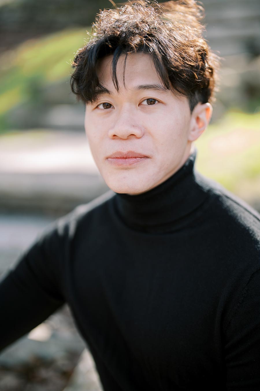 A portrait of a person wearing a black turtleneck, with a blurred outdoor background.