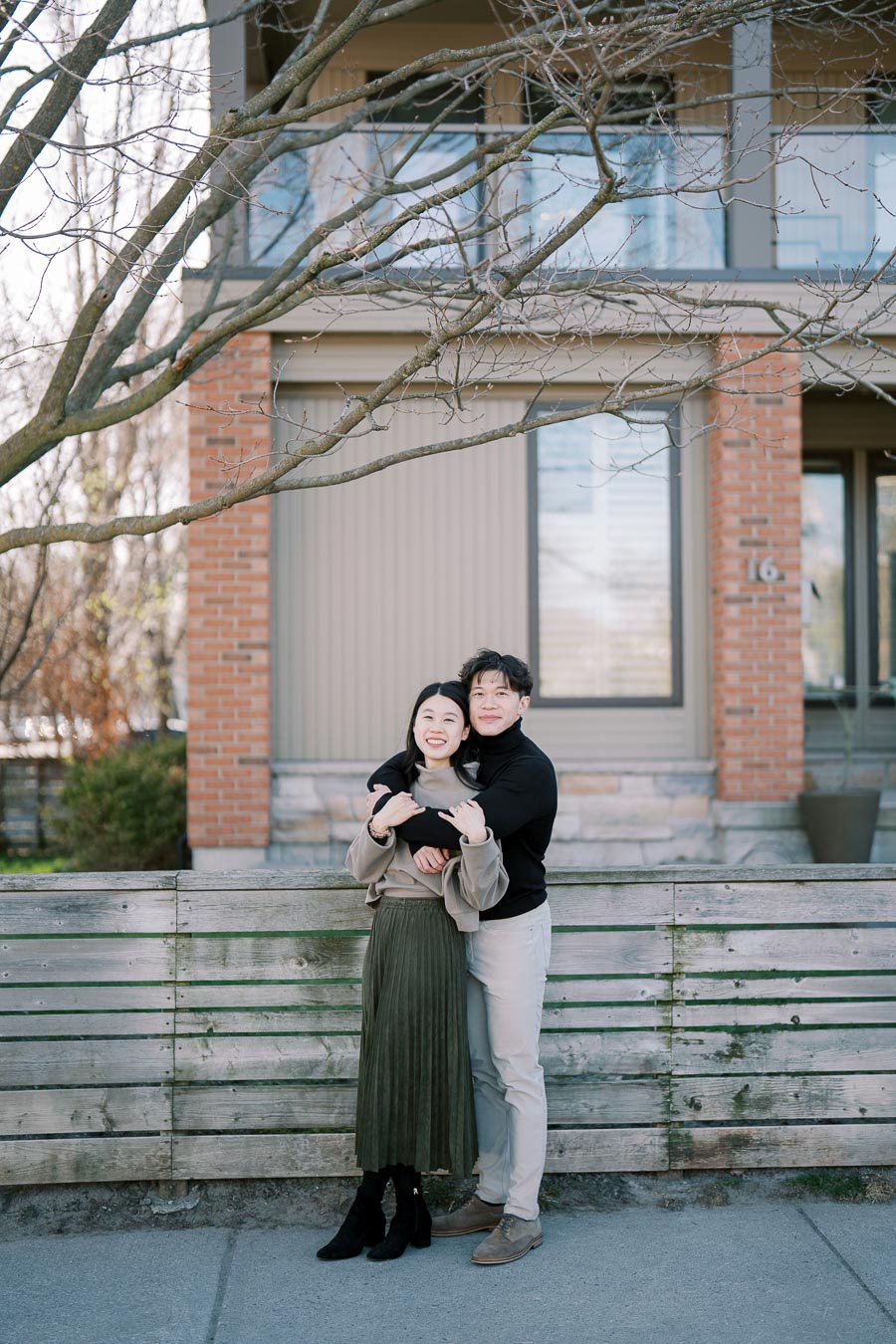A couple embraces in front of a modern house with a wooden fence and bare tree branches, enjoying a sunny day.