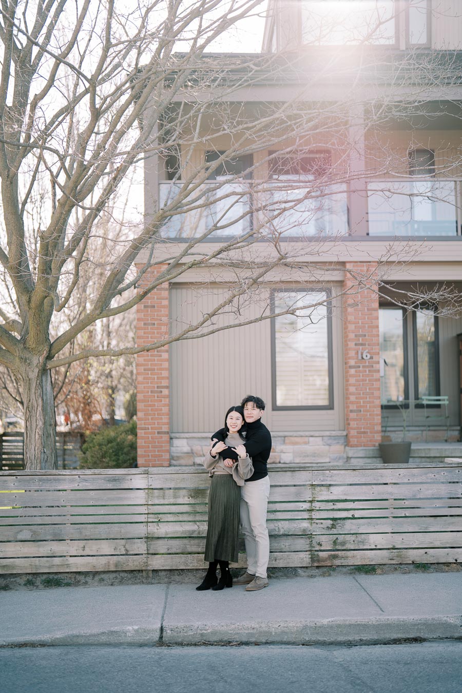 A couple embraces in front of a modern house with a wooden fence and bare tree branches, capturing a serene moment.