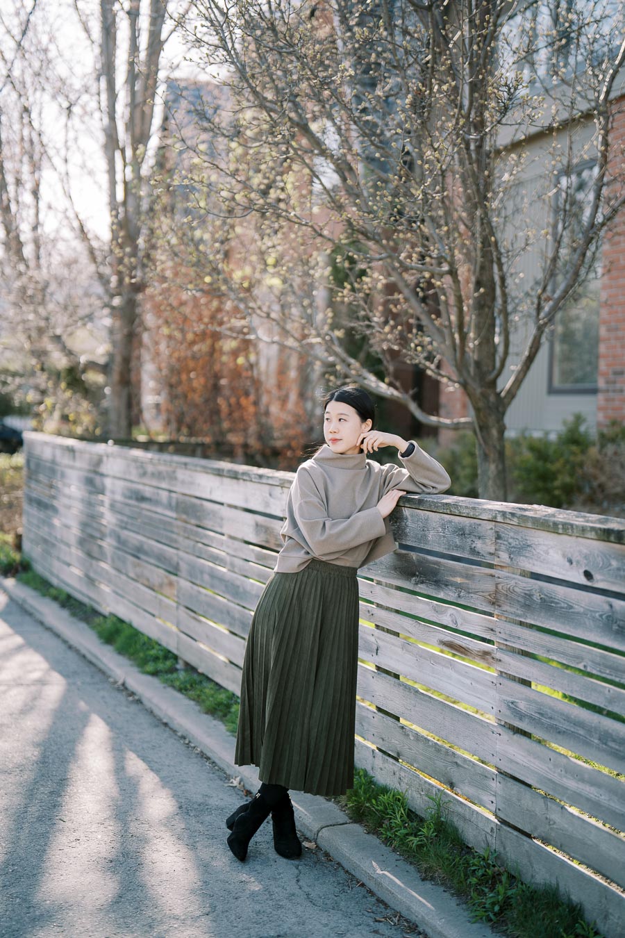 A woman in a casual outfit leans against a wooden fence, surrounded by early spring foliage and a house in the background.