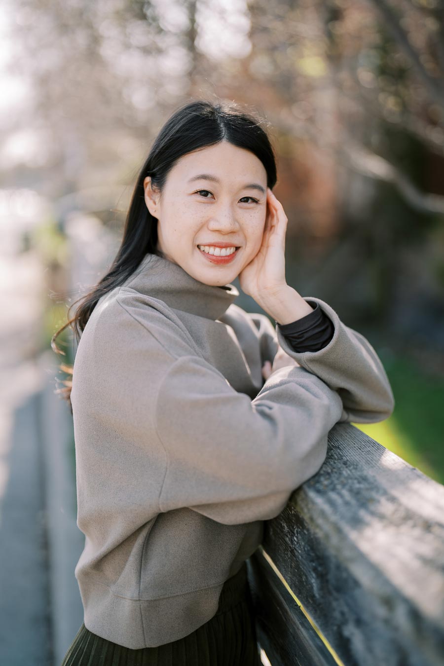 A woman in a beige sweater smiles while leaning on a wooden fence in a park setting.