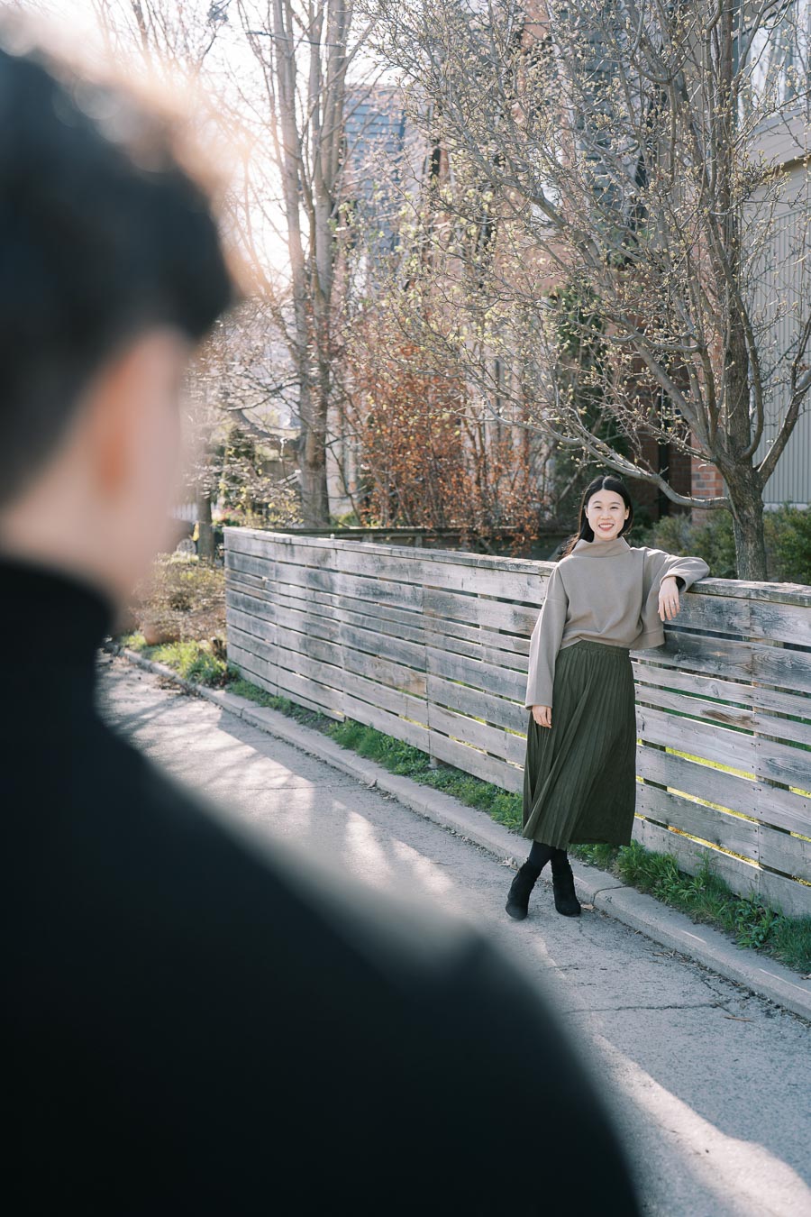 A woman poses by a wooden fence on a sunny day, with a blurred figure in the foreground.