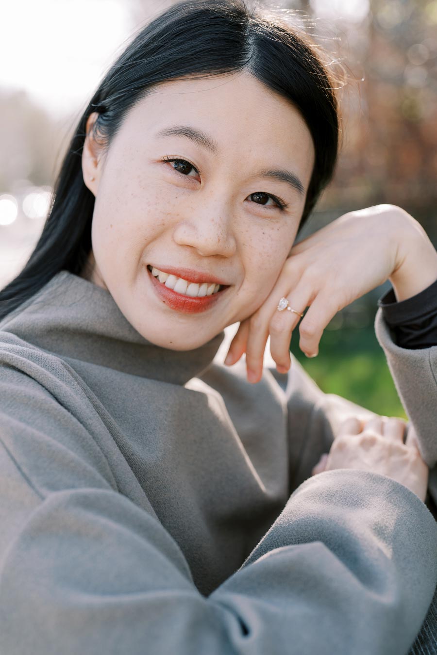 A woman smiling outdoors, wearing a gray coat and displaying a ring on her finger.