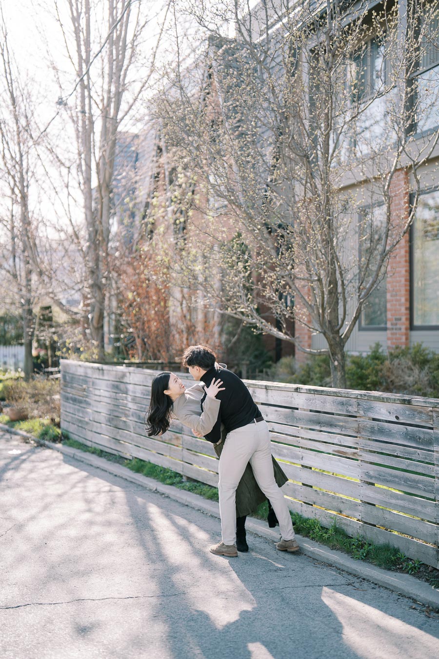 A couple shares a joyful moment on a sunny street, surrounded by budding trees and a wooden fence.