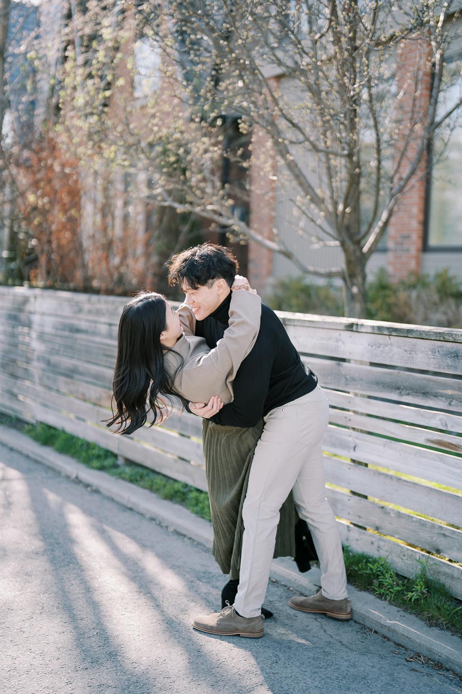A couple shares a joyful embrace on a sunny day, standing near a wooden fence with trees in the background.
