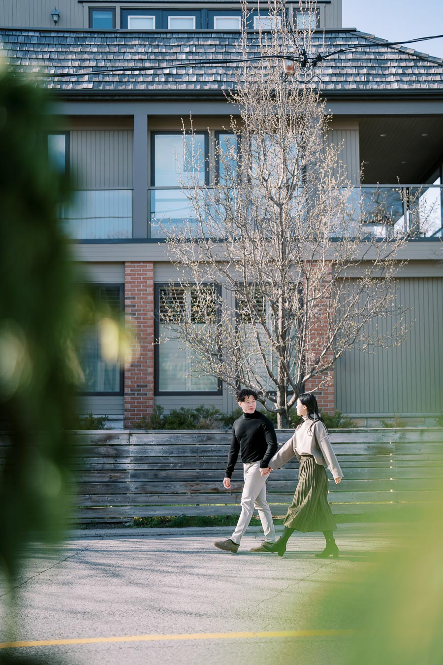 A couple strolls hand in hand past a modern house with a bare tree, enjoying a sunny day.