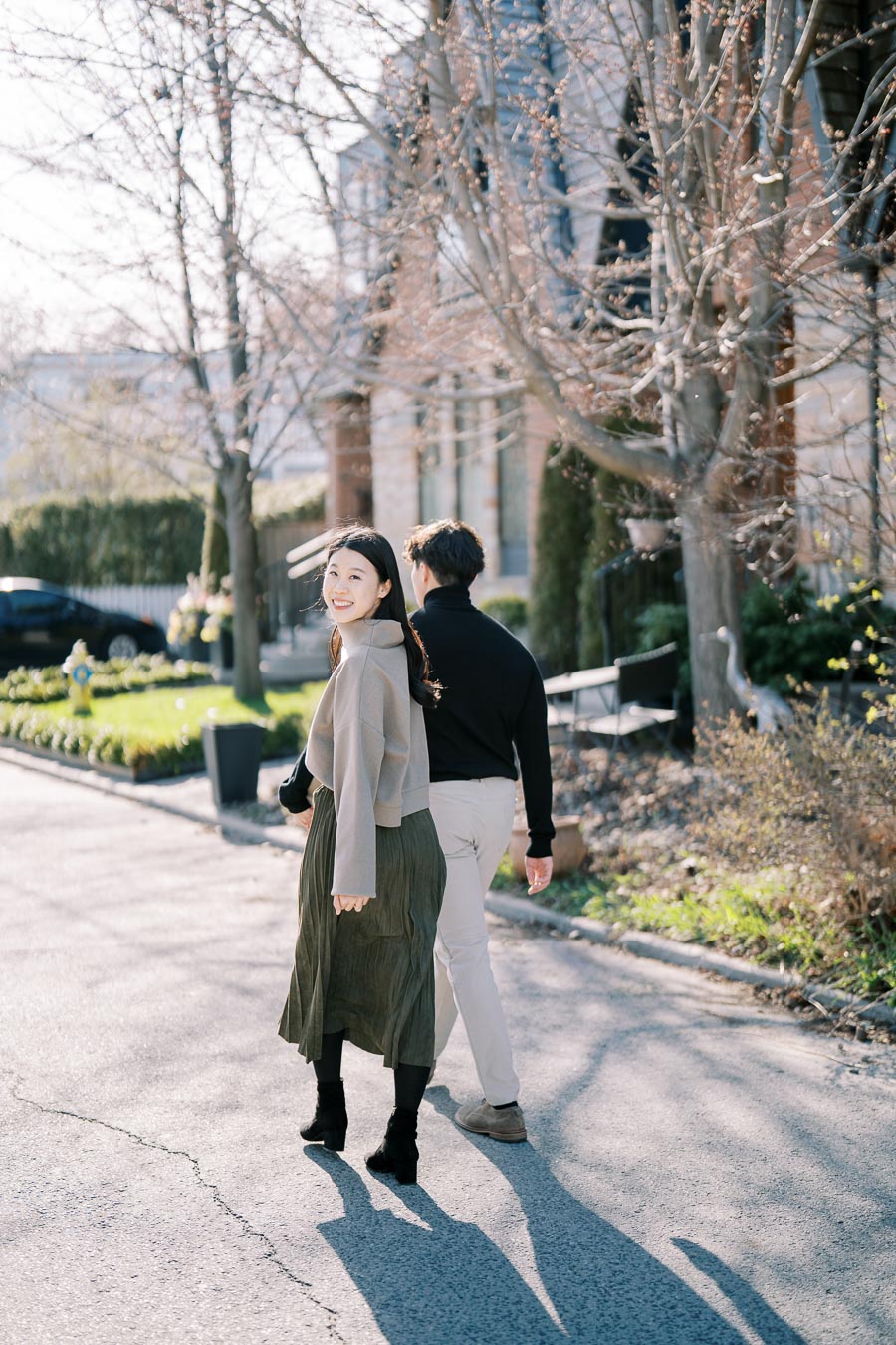 A couple enjoys a sunny walk on a quiet street lined with bare trees and charming houses.