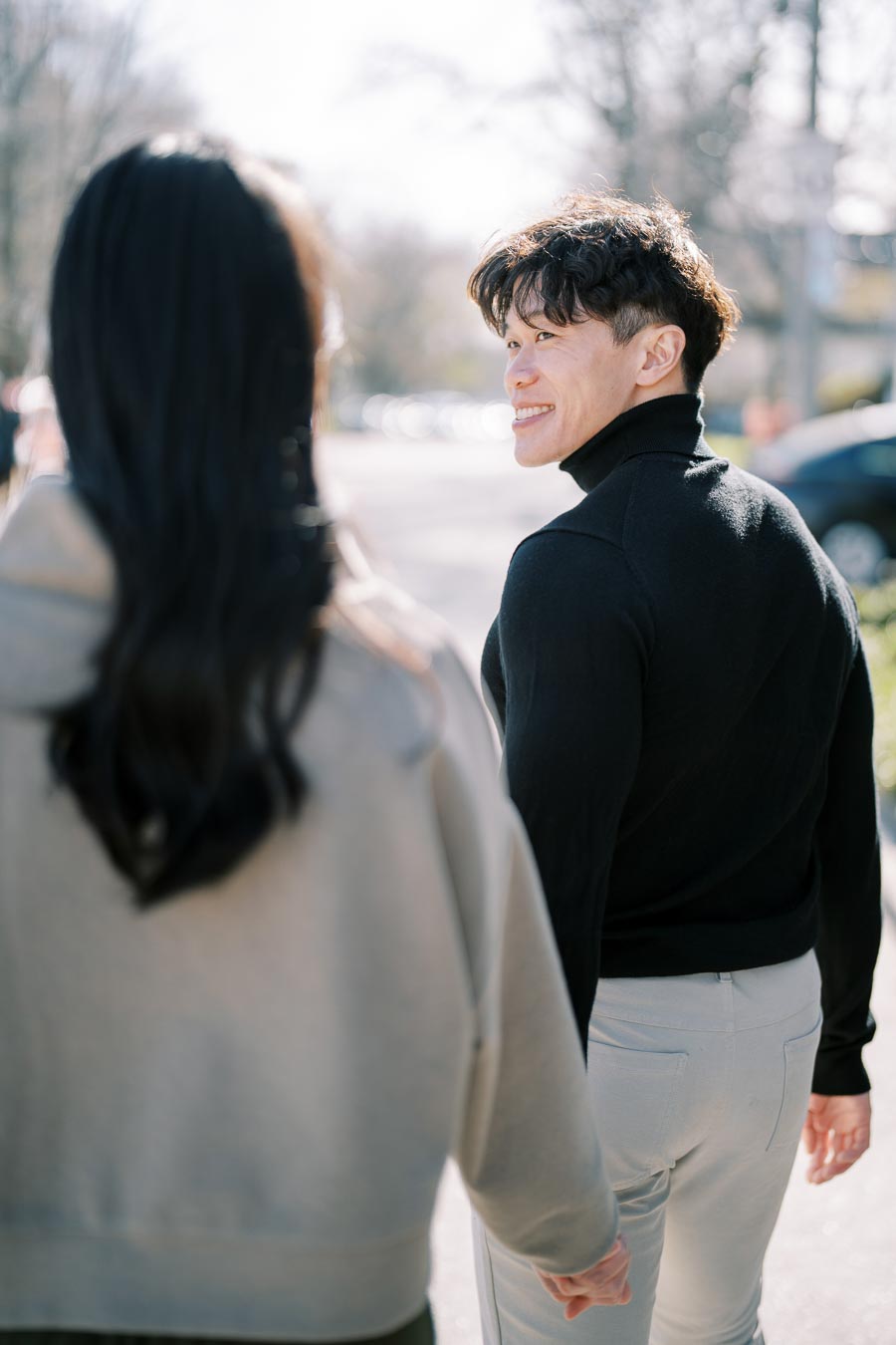 A couple enjoys a sunny day, walking hand in hand as the man looks back with a smile.