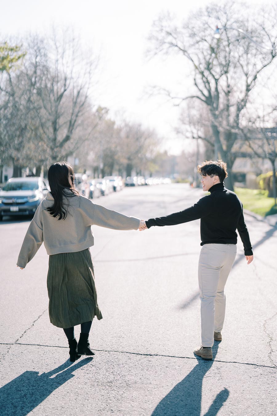 A couple walks hand in hand down a sunlit street, surrounded by bare trees and parked cars, enjoying a peaceful moment together.