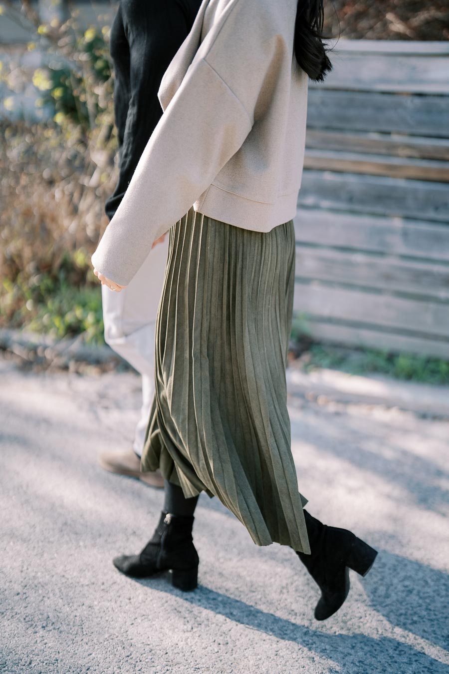 A close-up of two people walking, showcasing a beige sweater and green pleated skirt paired with black boots.