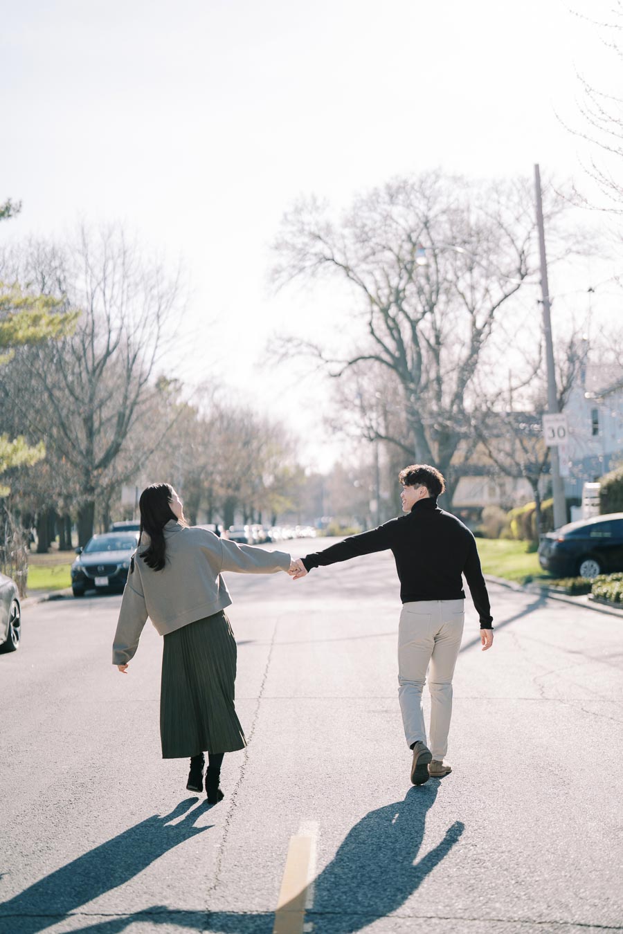 A couple joyfully walks hand in hand down a sunlit street, surrounded by bare trees and parked cars.