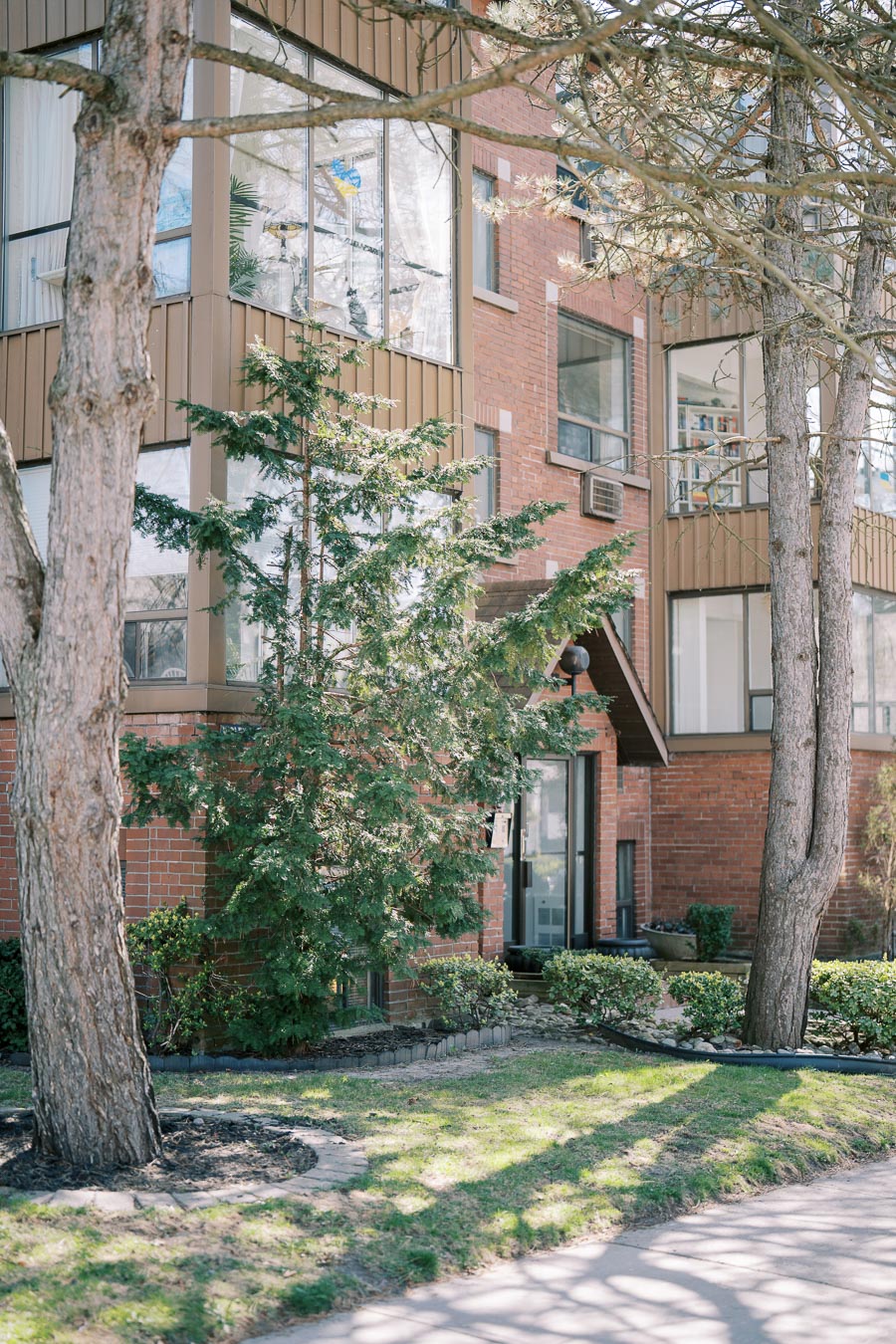 A brick apartment building with large windows is surrounded by trees and greenery, creating a serene residential environment.
