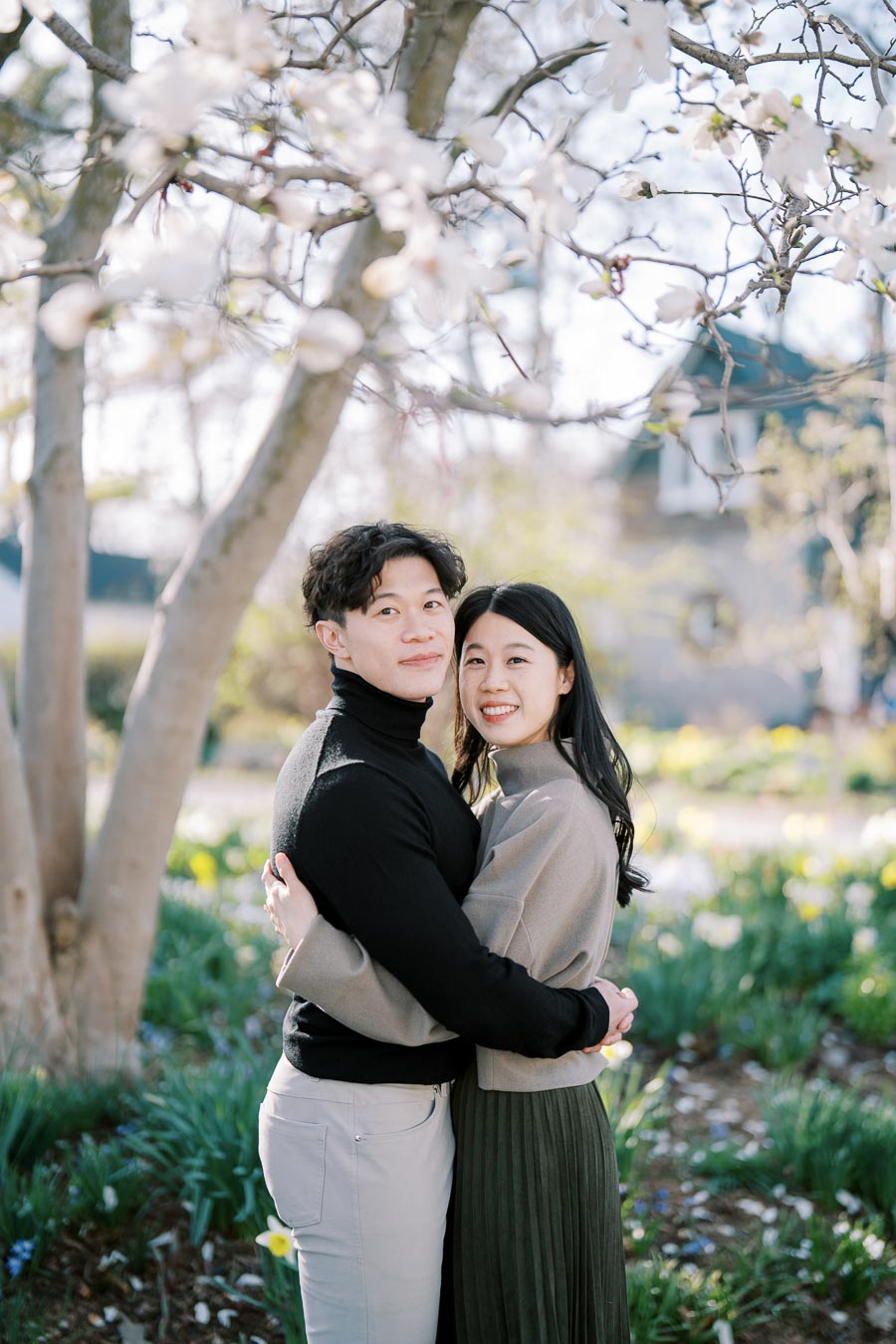 A couple embraces under a blooming tree in a garden, surrounded by spring flowers.