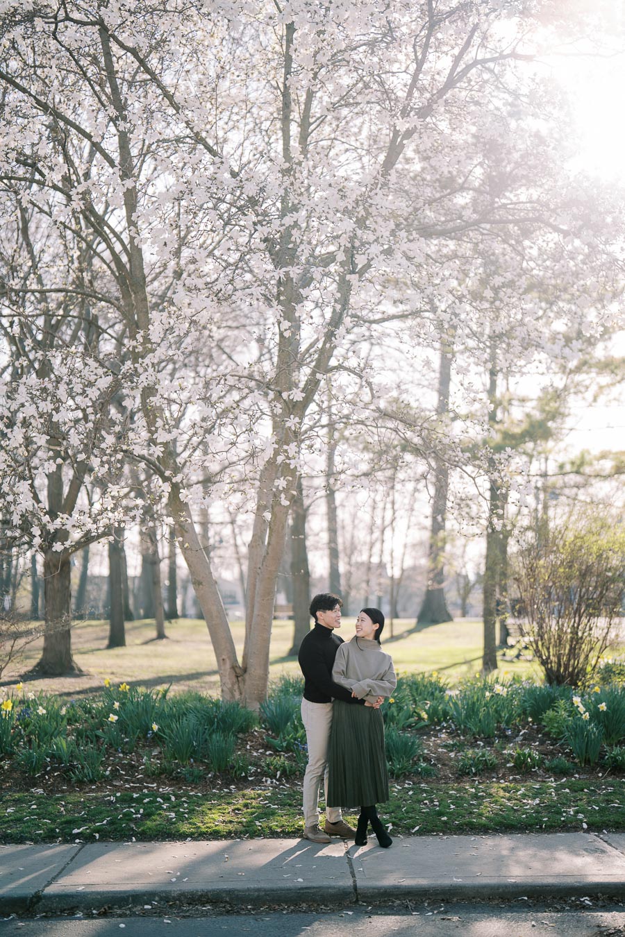 A couple stands under a blooming tree in a park, surrounded by spring flowers and sunlight filtering through the branches.