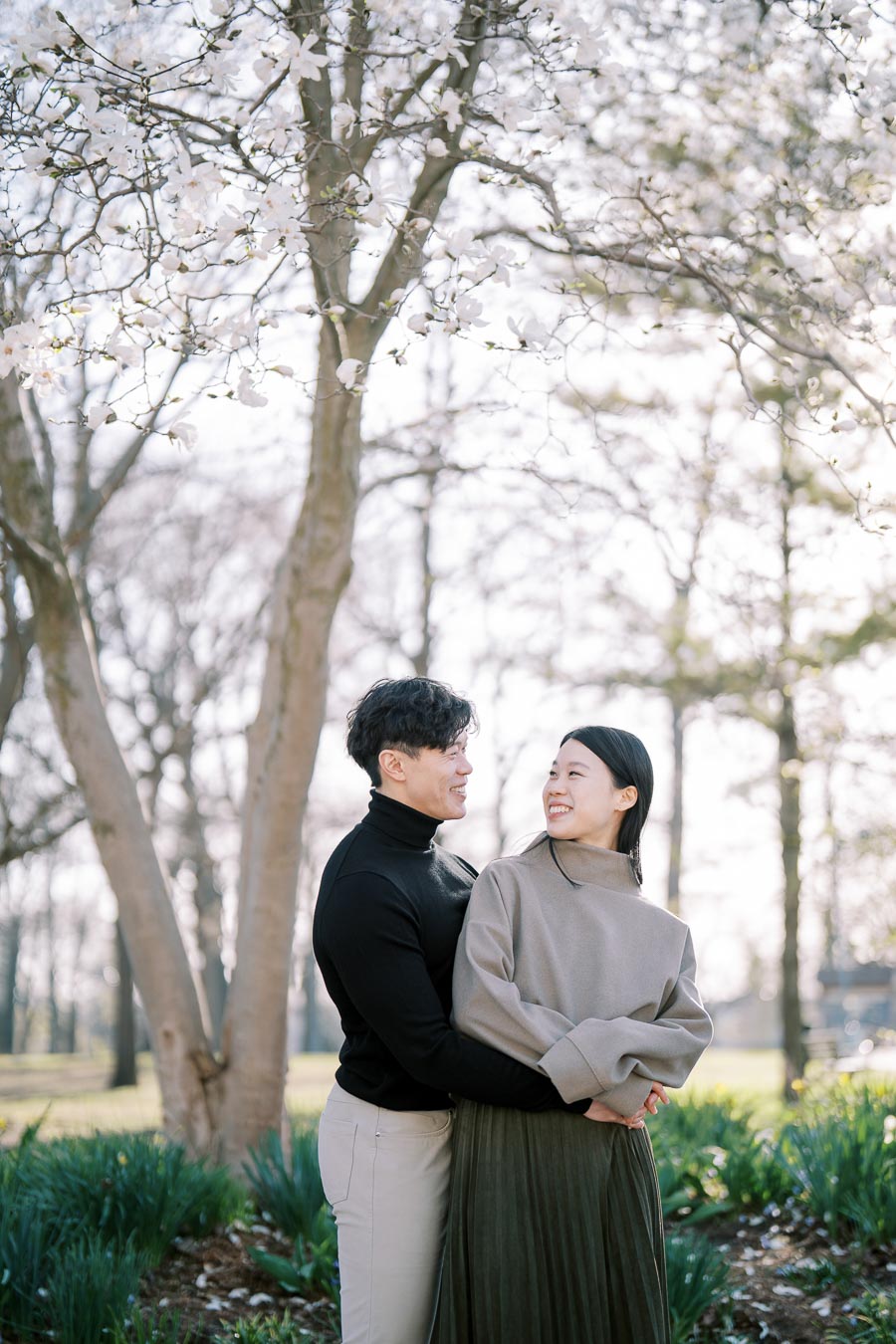 A couple embraces under a blooming tree in a park, surrounded by greenery and spring blossoms.